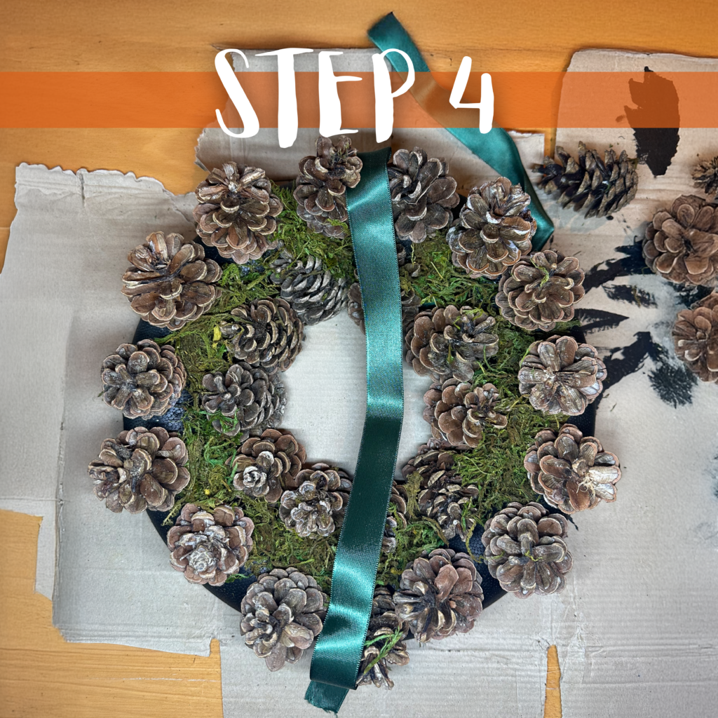 Das Band wird am fertigen Türkranz befestigt, indem es zwischen den Tannenzapfen durchgezogen und zugeknotet wird.